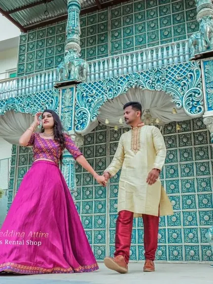 This image shows a couple in traditional attire, with the woman in a magenta skirt and embroidered top, and the man in a golden kurta set. The outfits are perfect for shoots at locations with architectural details.