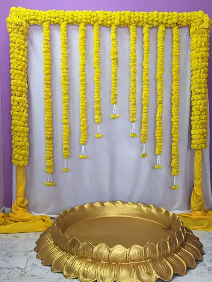 A simple and classic Haldi ceremony backdrop. The decor consists of a frame draped in white and yellow fabric with hanging marigold garlands.