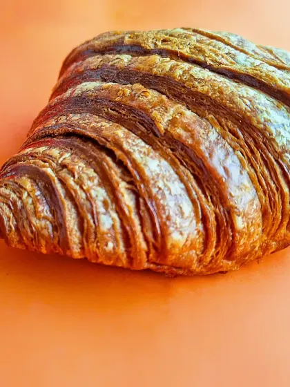 A simple, elegant shot of a single croissant against a solid orange background. This minimalist approach focuses entirely on the pastry's flaky layers and golden texture, perfect for packaging or clean ad campaigns.