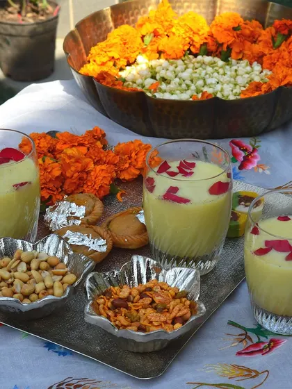 A festive Holi scene featuring our silver-plated tray and floral bowls, perfect for serving thandai and snacks. The hammered urli in the background adds a traditional touch with marigold flowers.