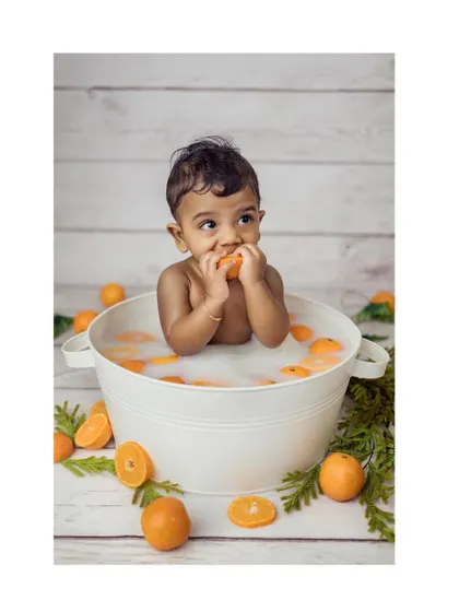 A delightful photo of a baby boy sitting in a miniature tub for a milk bath, surrounded by bright oranges. He is happily nibbling on an orange slice, creating a fun and vibrant image.