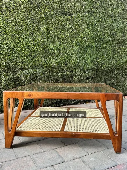 A square coffee table with a glass top, a teak wood frame, and a lower shelf made of hand-woven cane. This mid-century design is both elegant and functional.