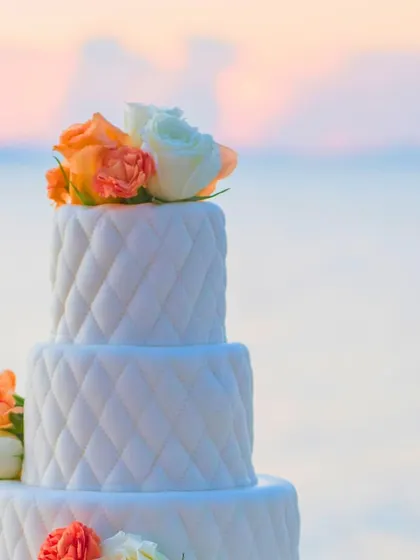 A beautiful three-tiered wedding cake with a quilted pattern, topped with fresh orange and white roses, set against a serene sunset backdrop. A sweet ending to a perfect day.