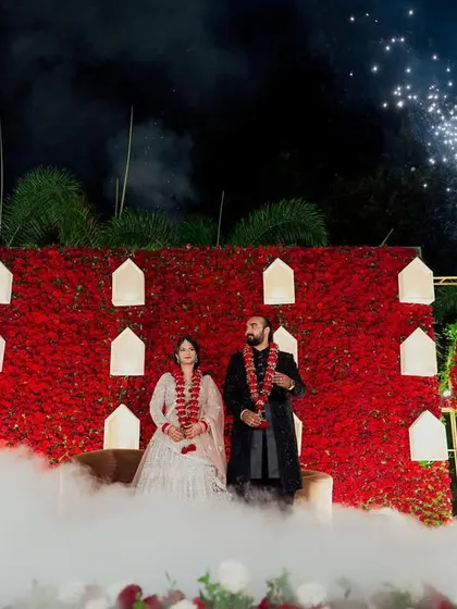 A dramatic stage for a memorable reception moment. The deep red floral wall is accented with illuminated niches, while cold pyro fireworks and low-lying fog create a theatrical and romantic atmosphere for the couple.
