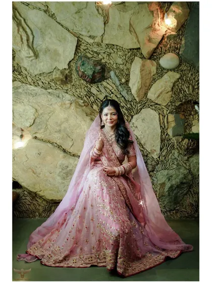 This full-length portrait showcases the bride's magnificent lehenga against a textured stone wall. The soft lighting and her gentle pose create an image of pure elegance.