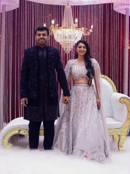 The couple posing on their reception stage, which features a dramatic backdrop of burgundy drapes and crystal curtains. A large chandelier adds a touch of opulence to the entire setup.