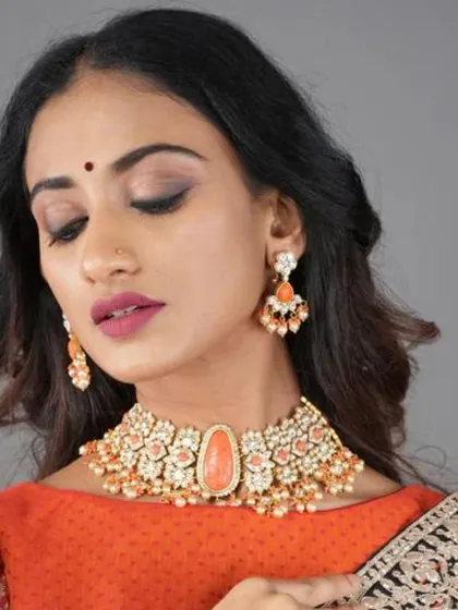 A model wears a stunning choker set featuring a large, peach-colored central stone surrounded by intricate kundan-style work and pearls. The matching drop earrings complete this festive ensemble.