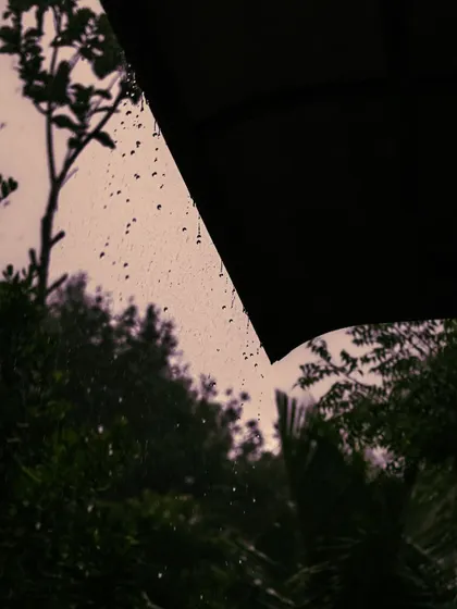 An artistic shot of rain seen from under an umbrella. The dark silhouette against the rainy sky creates a poetic and contemplative mood.