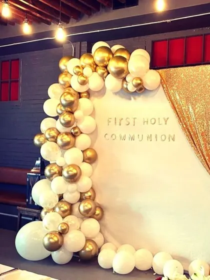 An elegant Holy Communion photo corner with a white backdrop and a gold sequin curtain. The scene is framed by a white and gold balloon garland.