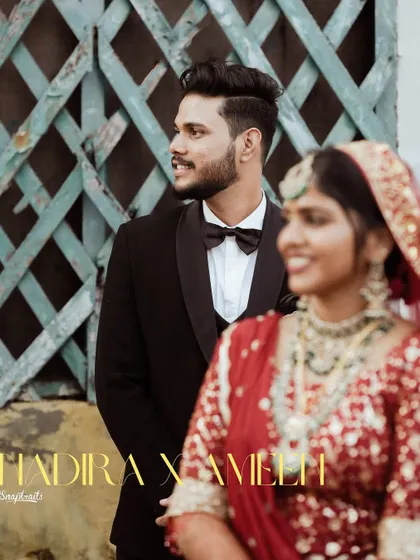 A stylish couple poses against a rustic, textured background, blending modern wedding attire with a touch of vintage charm.