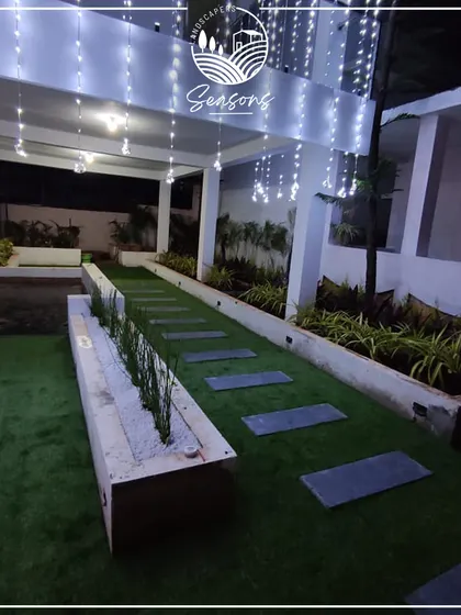A modern residential landscape featuring an artificial lawn, stone slab pathway, and a raised planter bed. The string lights add a magical touch, making the space perfect for evening relaxation.