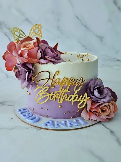 A beautiful floral cake in purple and white ombre. This chocolate truffle cake is adorned with artificial flowers and gold accents for an elegant birthday celebration.