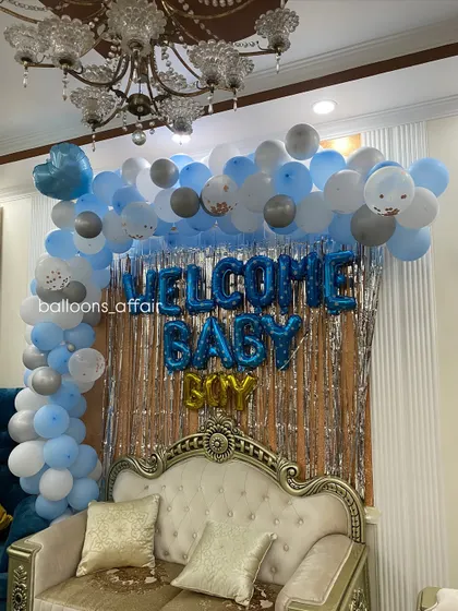 A 'Welcome Baby Boy' setup in a living room, featuring a balloon arch against a silver shimmer curtain and an elegant sofa.