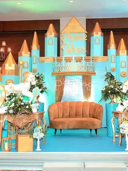 A regal stage setup for a prince's birthday, featuring a blue and gold castle backdrop, ornate crowns, and elegant floral arrangements. The central throne-like sofa provides the perfect spot for the birthday boy to preside over his celebration.