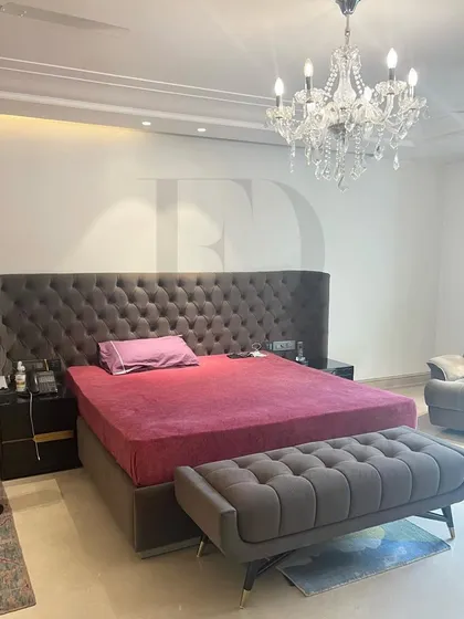A grand and opulent bedroom featuring a tall, button-tufted headboard in a dark grey fabric. The space is illuminated by a stunning crystal chandelier, adding a touch of classic glamour.