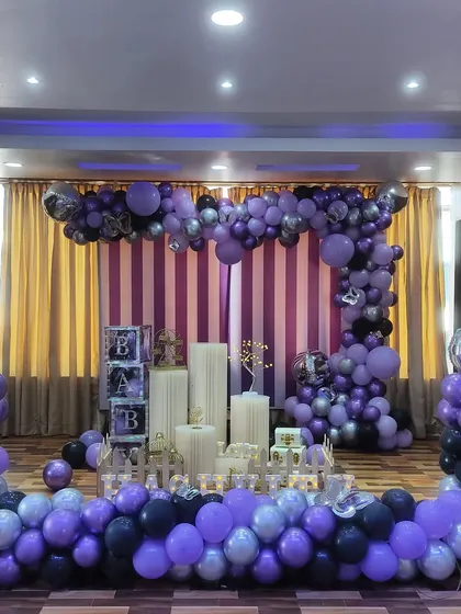 A grand naming ceremony setup in a function hall. The stage is decorated with a large balloon arrangement in purple, black, and silver, with two large disco balls for a fun, celebratory touch.