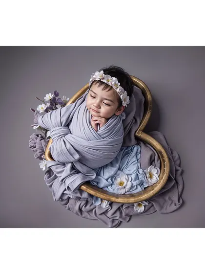 A sleeping baby in a heart-shaped prop, swaddled in a calming grey-blue wrap with delicate floral accents.