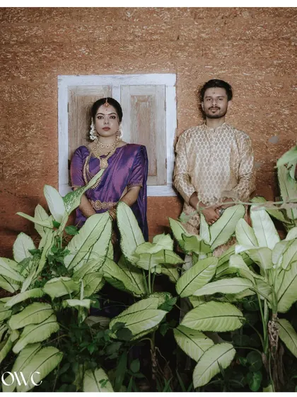 A formal portrait of a couple in striking traditional attire, posed against a rustic wall with lush green plants.