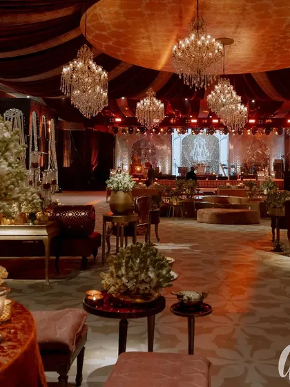A wide view of the reception hall, blending traditional Indian opulence with modern luxury. A color palette of deep red and gold is accented with ivory florals, while dozens of chandeliers create a majestic atmosphere.