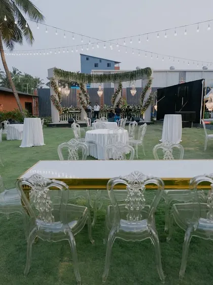 An elegant outdoor reception setup on a lush green lawn. We used a mix of round and rectangular tables with modern transparent chairs, all under beautiful string lighting, to create a sophisticated dining experience.