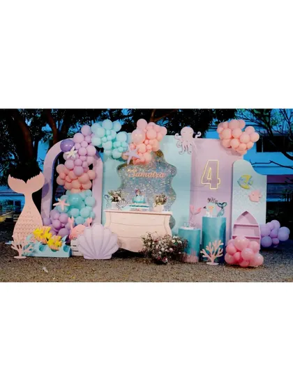 A wide shot of an outdoor mermaid party for a 4th birthday. The setup includes multiple backdrops, a large mermaid tail, and seashell props in a beautiful pastel palette.