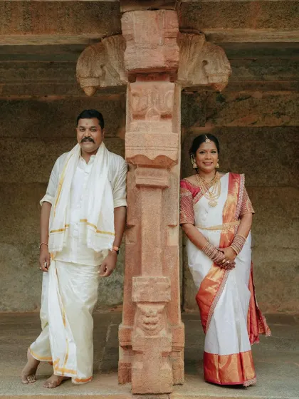A classic portrait of a couple in traditional South Indian wedding attire. They are posed against a carved stone pillar, which adds a touch of heritage and cultural significance to the photo.