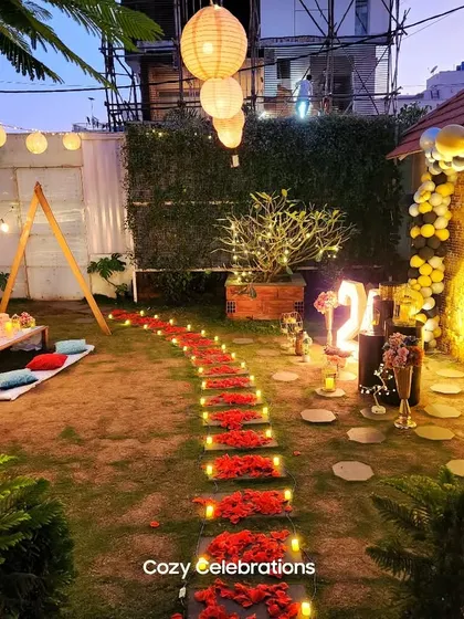 A wide-angle view of a romantic garden setup, featuring a candle and flower-petal pathway leading to a decorated arch and picnic area.