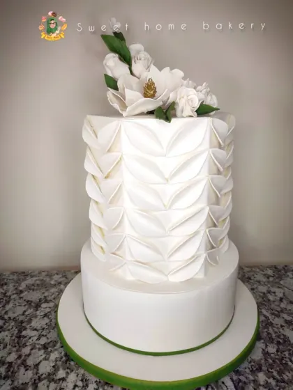 This two-tiered white wedding cake features a modern, geometric folded fondant technique on the top tier. It's complemented by a beautiful spray of handcrafted white sugar magnolias, creating a design that is both contemporary and timeless.