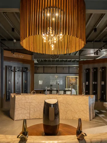 Inside the Satyam showroom, a large, custom chandelier made of wooden slats hangs over a marble-clad display counter. This central feature adds a touch of warmth and sophistication to the industrial interior.