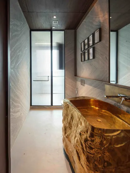 The powder room adjacent to the family lounge features a glamorous, sculptural brass washbasin console. The walls are clad in grey limestone, and the space is adorned with an evocative art piece titled 'Memories of an Old Tree'.