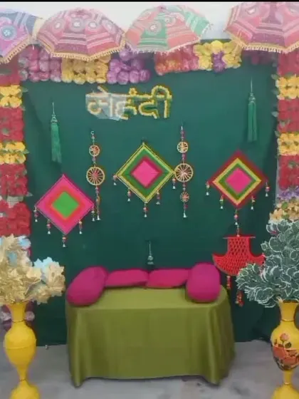 A Mehendi backdrop with a dark green curtain, decorated with colorful umbrellas, hanging props, and floral arrangements on the sides.