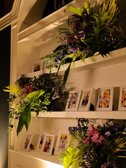 A closer view of the 'Little Big Moment' photo shelf. We integrated cascading floral arrangements among the family pictures, blending personal memories with our bespoke event design.