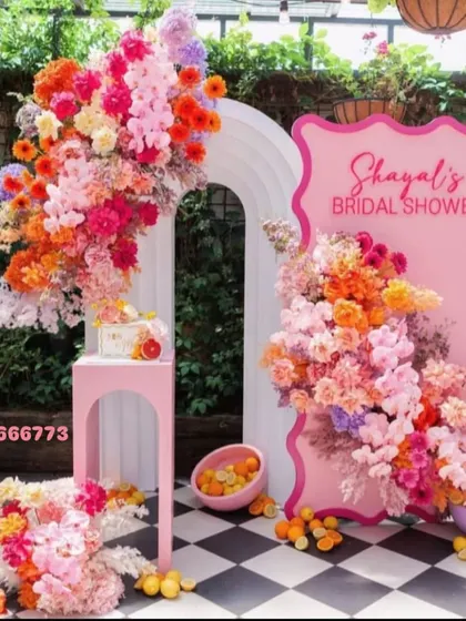 A modern bridal shower setup with a custom pink backdrop and a white arch. The decor is built around vibrant orange and pink fresh flower arrangements.