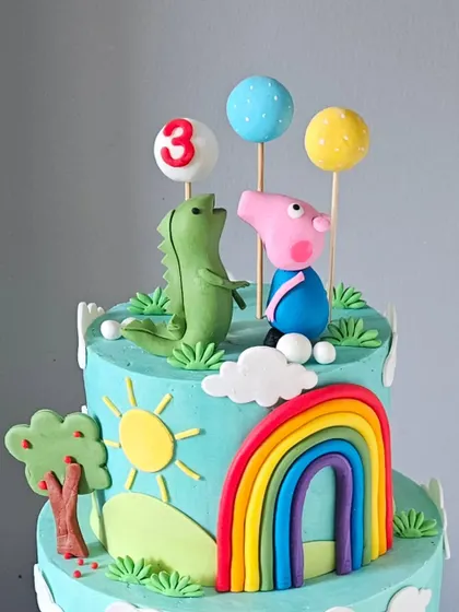 A close-up of the top of the Peppa Pig cake, showing Peppa and George's dinosaur. The bright colors and cheerful details like the sun and rainbow are all crafted from fondant.