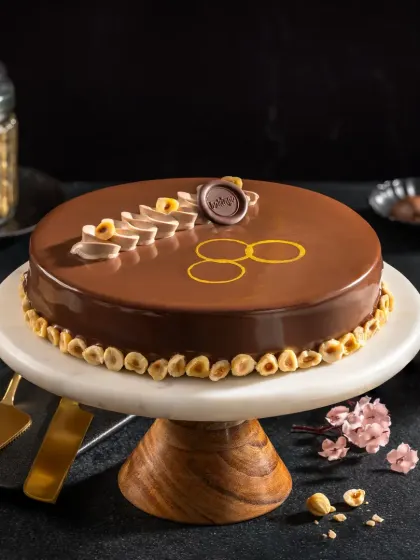 A glossy hazelnut chocolate cake with delicate piping and gold leaf accents, styled for a premium bakery's promotional materials.