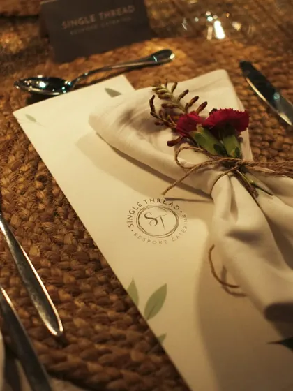A close-up of a place setting, featuring a custom menu with my logo, a floral-tied napkin, and elegant cutlery on a woven mat.