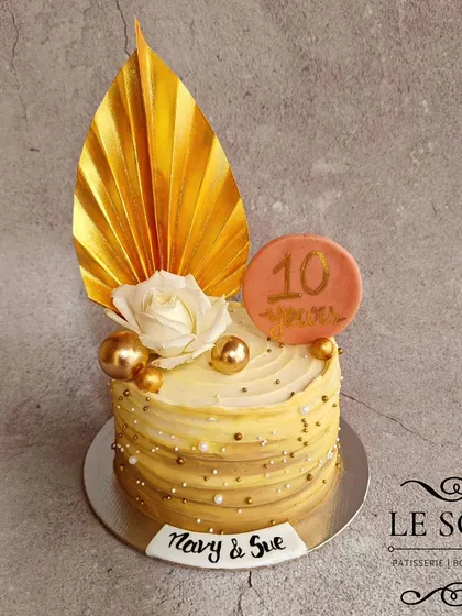 This anniversary cake features a beautiful striped buttercream effect in shades of gold and white. It's decorated with a handmade golden palm leaf, a fresh white rose, and delicate pearl sprinkles.