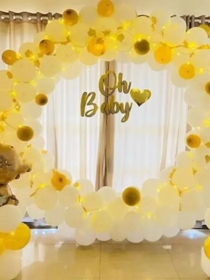 A simple and elegant "Oh Baby" shower setup with a full ring of white and yellow balloons. Fairy lights woven into the balloons and baby-themed foil balloons complete this cheerful look.