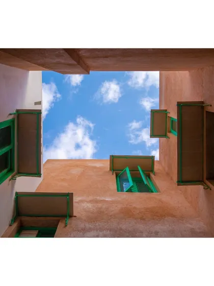 A view looking up from another courtyard, this one with earthy-toned walls and bright green window frames. The use of color and texture helps define different spaces and creates a visually rich environment.