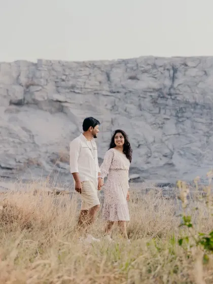 A candid shot of a couple walking through a field of tall, dry grass, with a massive rock formation in the background.