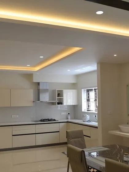This spacious open-plan area showcases a minimalist all-white modular kitchen that blends seamlessly with the dining space. The design is clean and bright, with layered ceiling lights providing ample illumination for both cooking and dining.