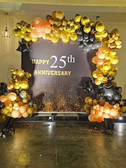 A stylish 25th-anniversary backdrop. The decoration uses a black, gold, and peach balloon arch to frame a custom "Happy 25th Anniversary" sign.