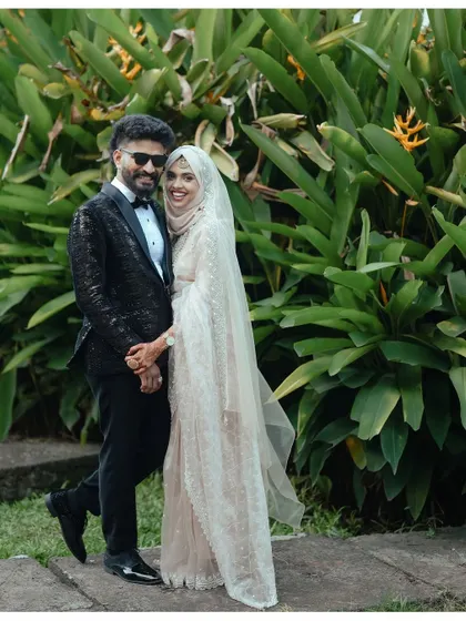 A classic wedding portrait of a Muslim couple, beautifully framed by lush green foliage. Their coordinated outfits and happy smiles make this a timeless keepsake.