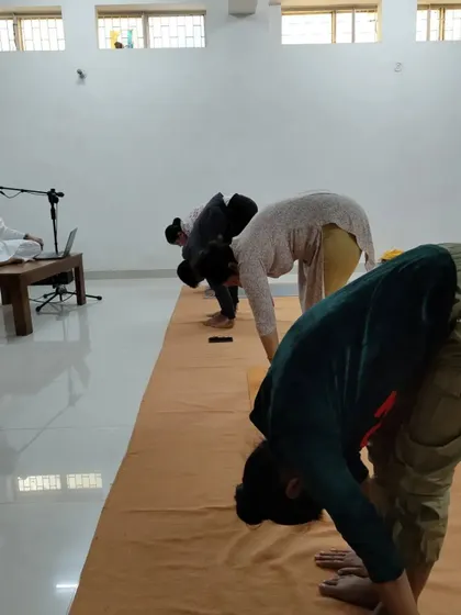 The teacher observes as participants practice a forward bend, ensuring correct form and alignment.