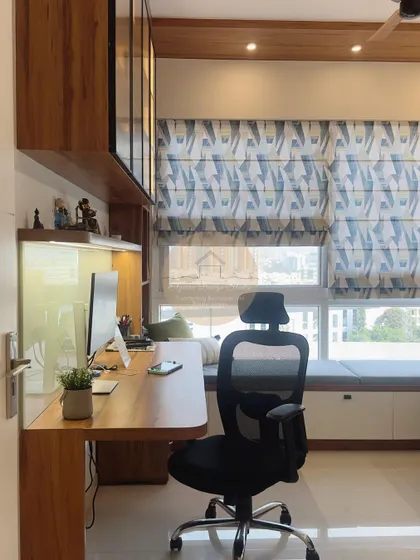 A cozy and functional study cum office space. This design features a long wooden desk, ample storage with overhead cabinets and a window-side bench, and beautiful patterned roman blinds that control natural light.