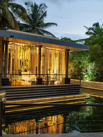 A stunning evening view of a modern mandap set by the water. The structure is lit from within, creating a golden glow that reflects beautifully on the pond, making for a truly magical wedding venue.