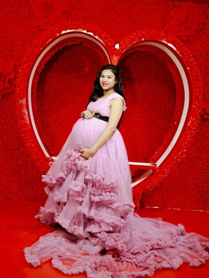 A beautiful solo shot of an expectant mother in a lavender ruffled gown, cradling her baby bump in front of a red floral heart backdrop.