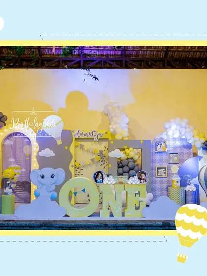 A cheerful and whimsical first birthday celebration with a hot air balloon and baby elephant theme. The complete stage setup features large 'ONE' block letters, custom backdrops, and a soft color palette of yellow, grey, and blue.