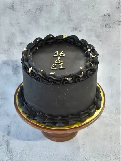 The top view of the black ganache cake, showing the "16 & 21" inscription for a joint sibling birthday, with delicate gold leaf accents on the piped border.