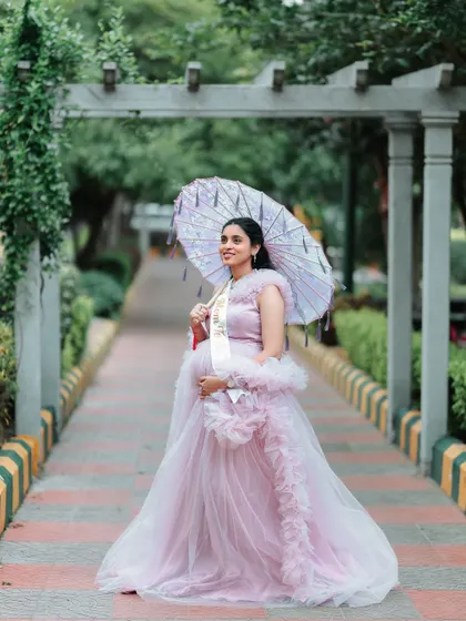 A full-length shot of the mom-to-be looking radiant. The parasol adds a vintage charm to the overall look, paired with the ruffled pink maternity gown.